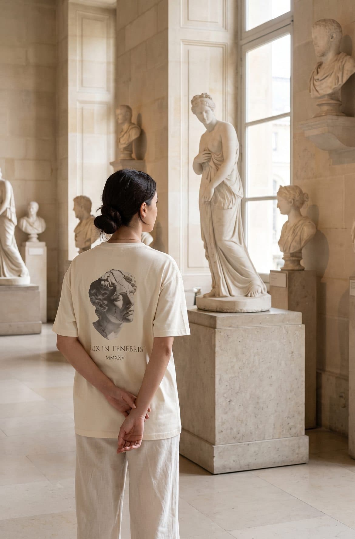 A woman in the Lux in Tenebris tee, seen from behind, studies a marble statue in a museum gallery